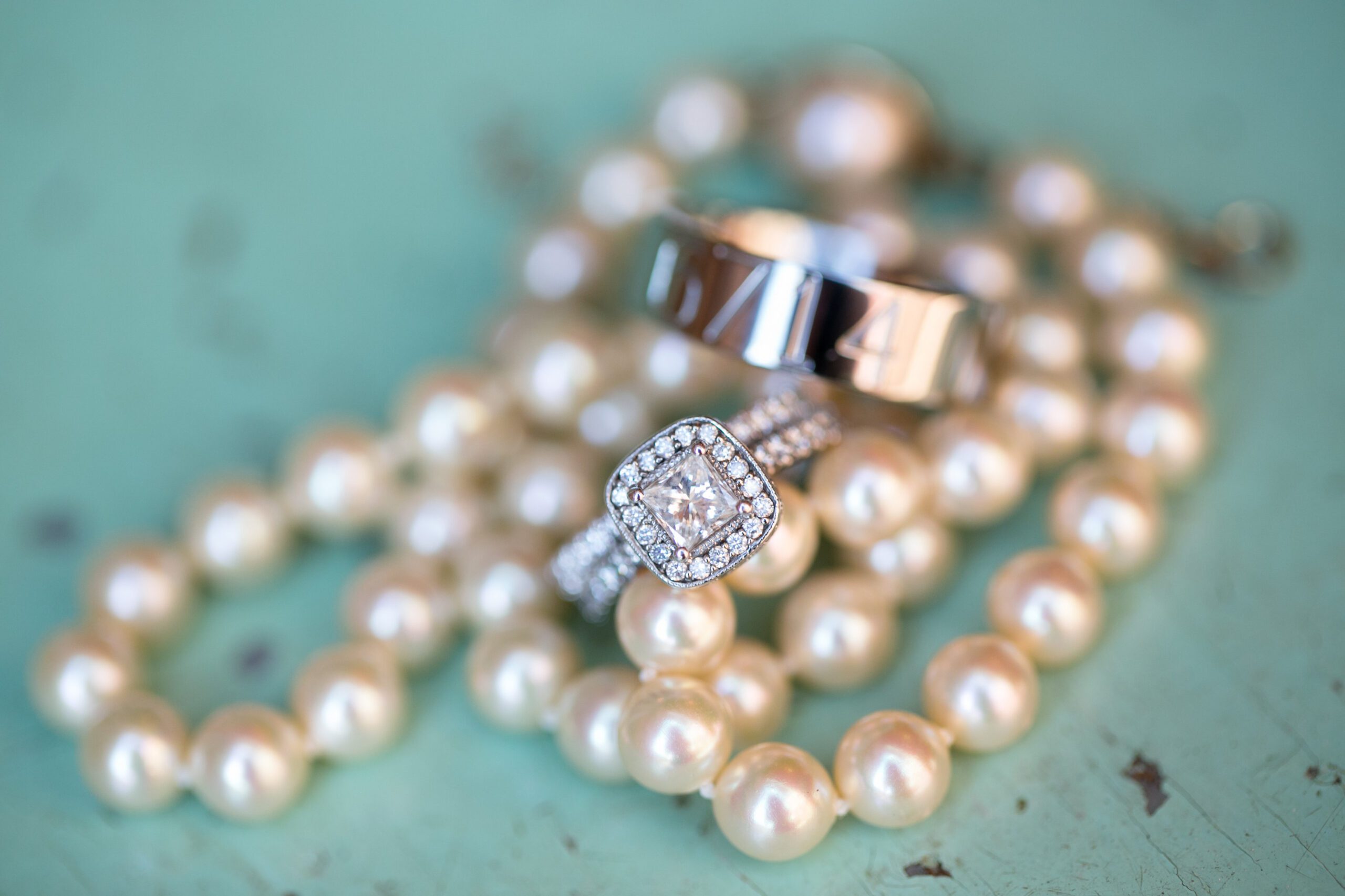 Close-up of a jewelry arrangement on a light teal surface. Featured are a string of pearls, a diamond-encrusted engagement ring with a square center stone, and a sleek wedding band. Captured by a Virginia Wedding Photographer, the pearls and sparkling diamonds create a luxurious and elegant display.