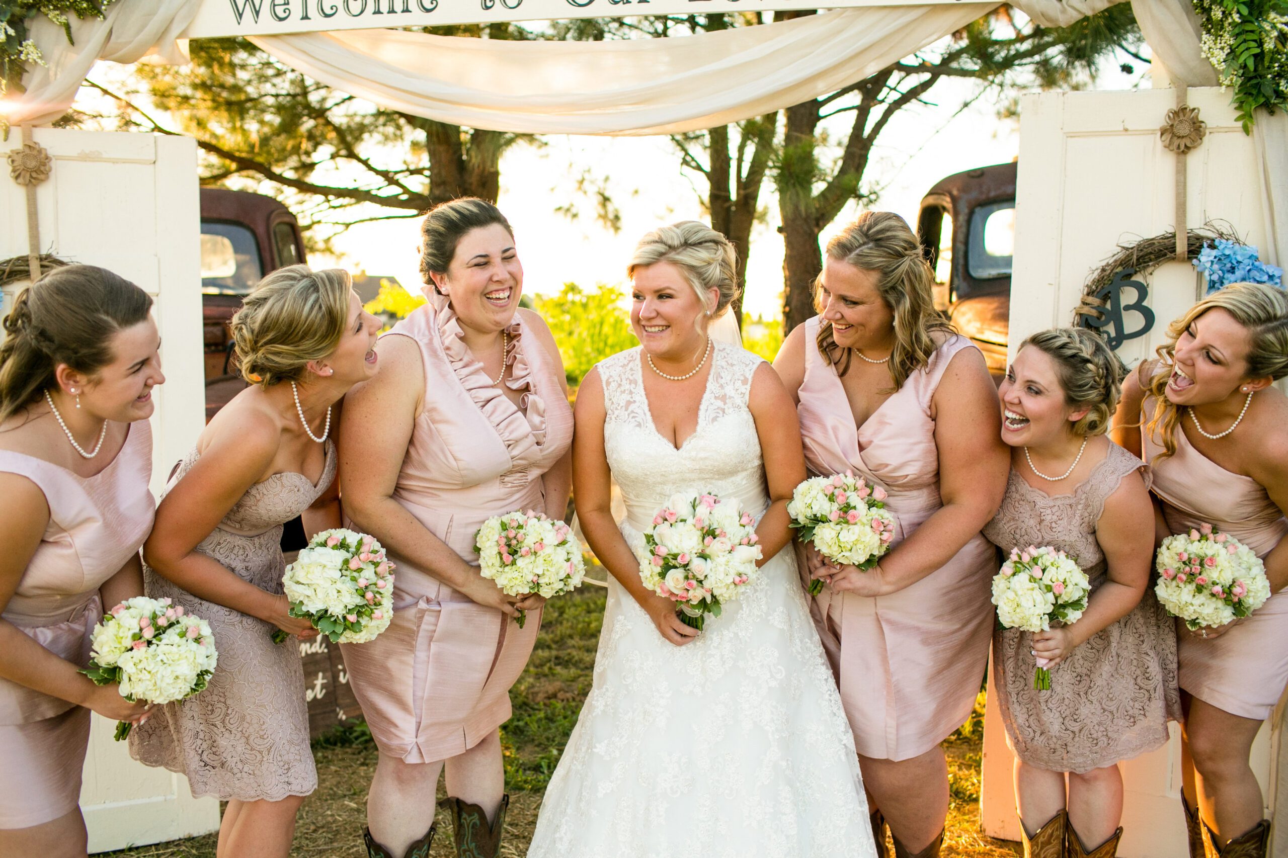 A bride in a white gown and six bridesmaids laugh together outdoors under a decorated archway. The bridesmaids wear light pink dresses, hold floral bouquets, and some wear cowboy boots. Trees and vintage trucks are visible in the background, beautifully captured by a Virginia Wedding Photographer.