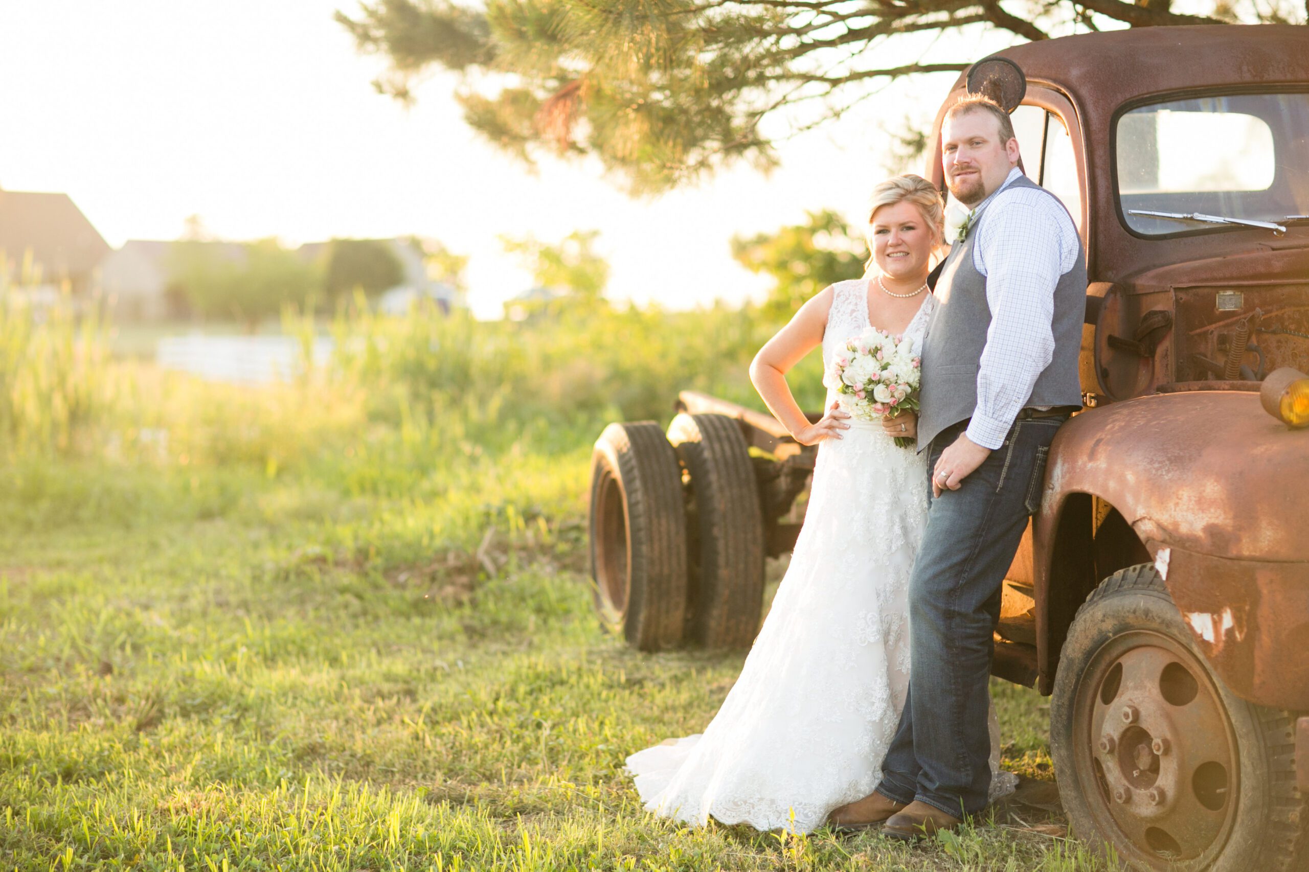 A bride and groom stand next to a rusted vintage truck in a sunlit field. The bride wears a white lace dress, holding a bouquet of flowers, while the groom is dressed in a blue shirt, gray vest, and jeans. They smile happily, posing for a photo under a tree captured by their Virginia wedding photographer.