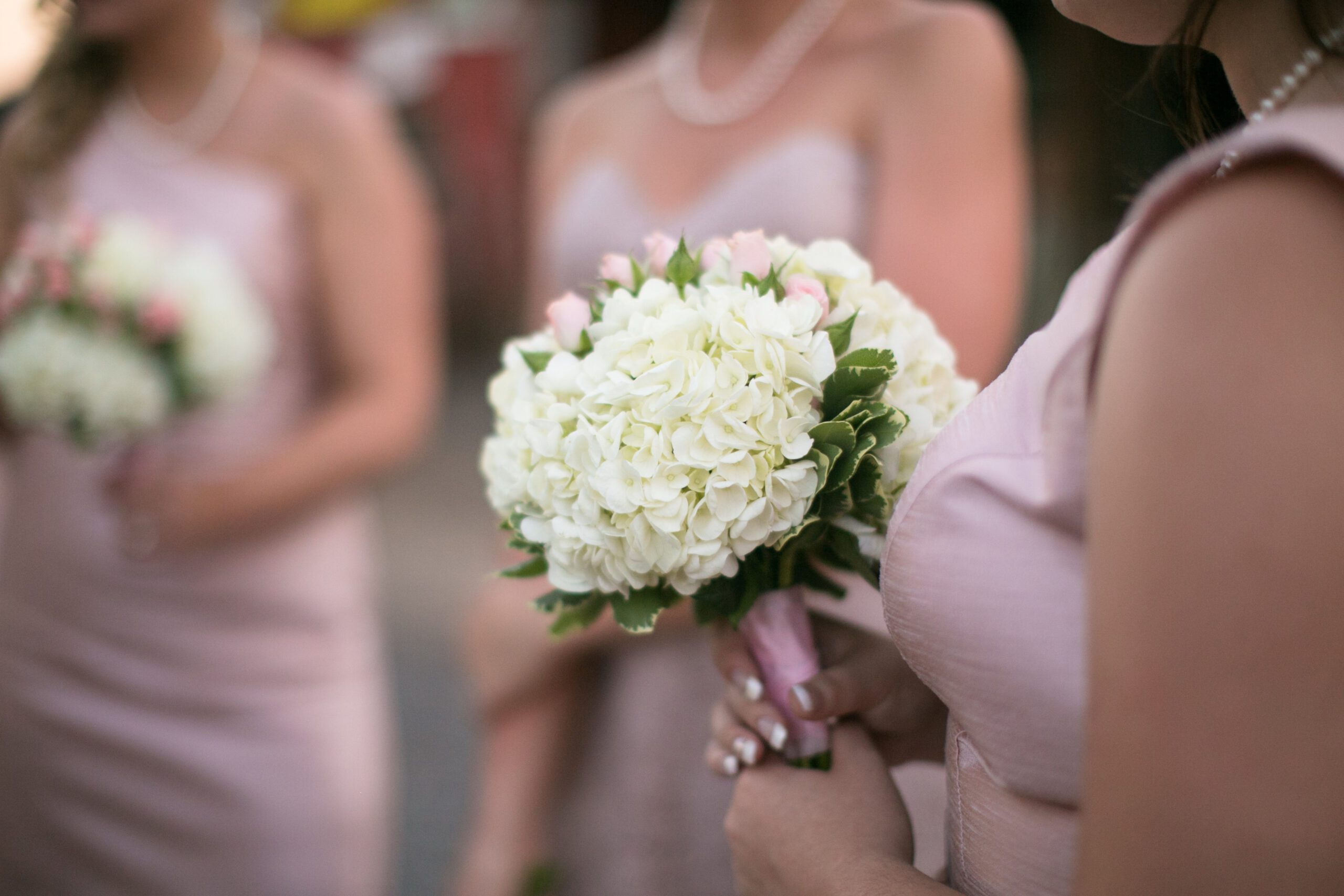 Close-up of bridesmaids in light pink dresses holding bouquets of white and pink flowers. The focus is on one bridesmaid's bouquet, showing detailed petals and green leaves, while two other bridesmaids with similar bouquets are visible in the background. Captured by a Virginia Wedding Photographer.