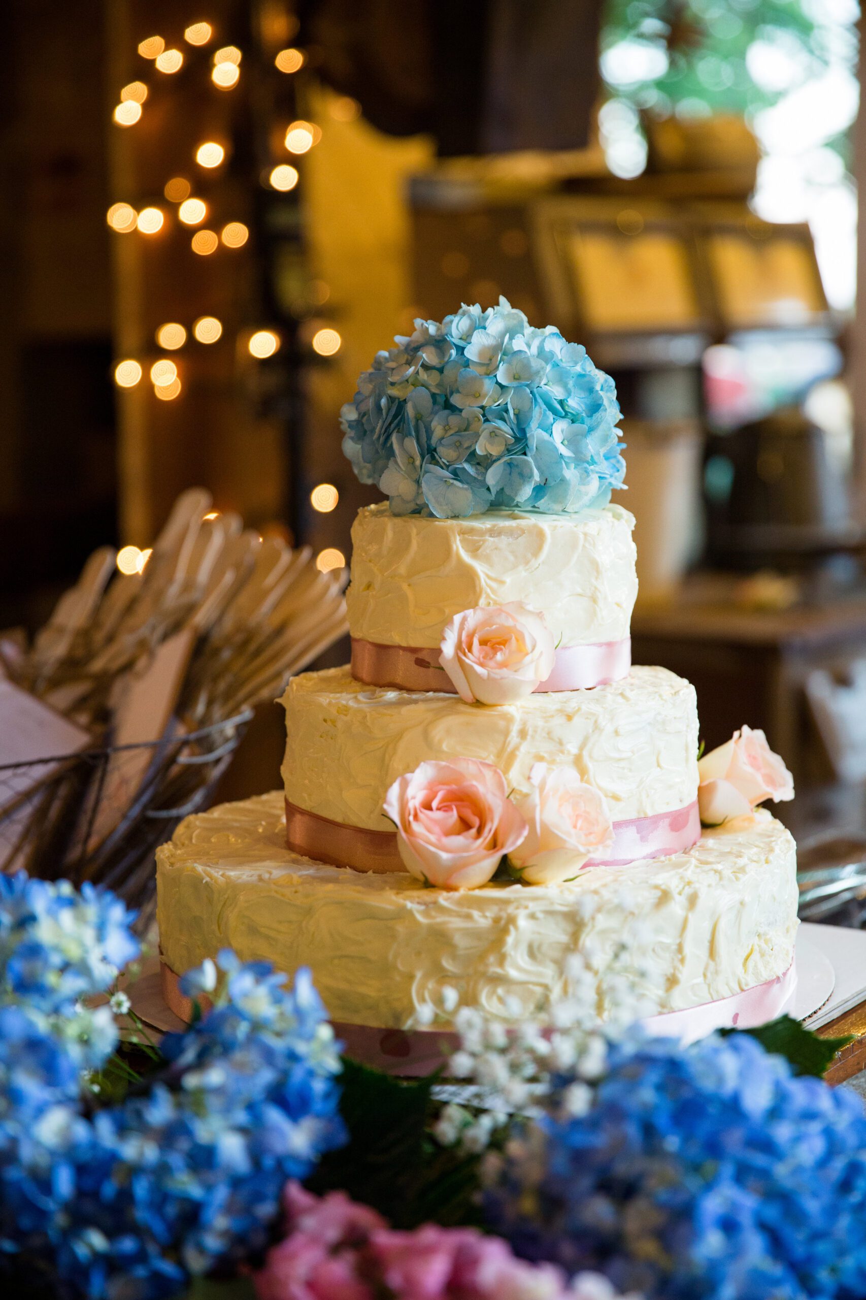 A three-tiered wedding cake with rustic, textured white icing is decorated with light pink roses and a bouquet of blue hydrangeas on top. A pink ribbon wraps around each layer, captured beautifully by a Virginia Wedding Photographer. The background features soft, ambient string lights and a blurred indoor setting.