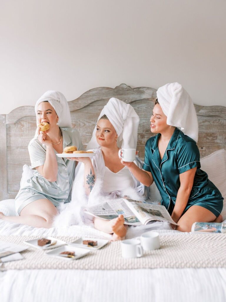 Three women in pajamas and towel turbans sit on a bed enjoying a weekend getaway breakfast. One woman holds a donut, another holds a plate with pastries and a drink, and the third has a cup of coffee. Newspapers and a breakfast spread are scattered around them. They are smiling and relaxed.