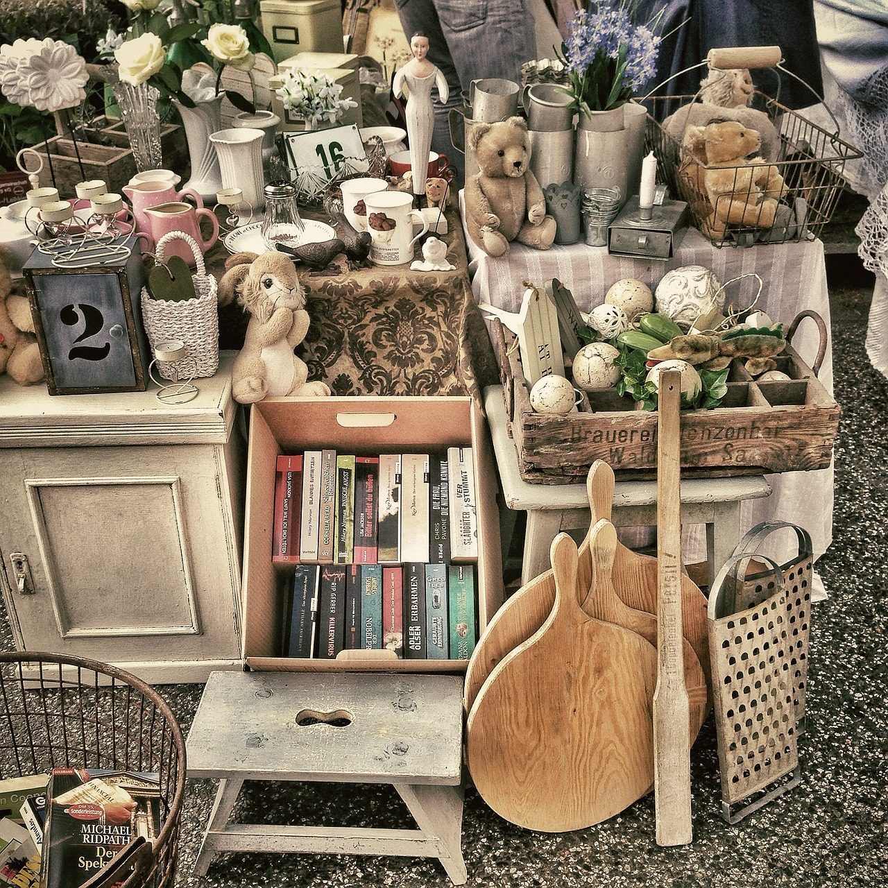 A collection of vintage items and books displayed at an outdoor market. The assortment includes stuffed animals, wooden cutting boards, ceramic dishes, a small cabinet, decorative figurines, and a variety of books organized in a cardboard box.