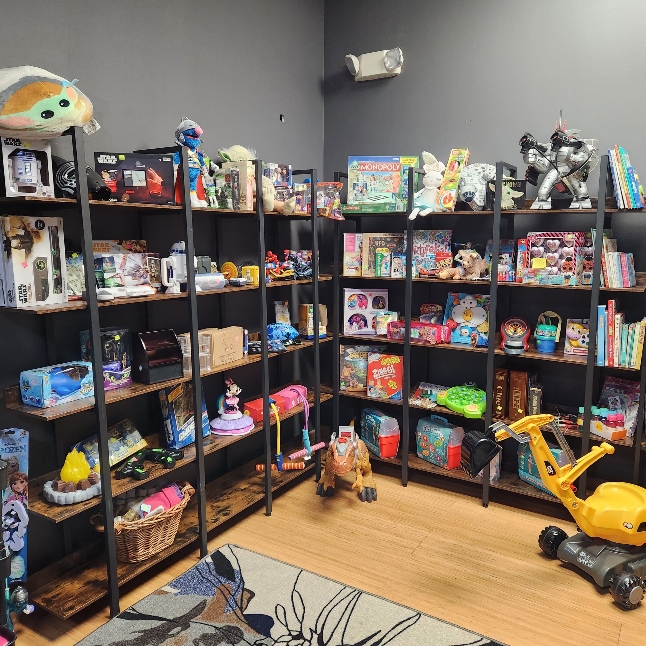 A room with dark walls and wooden flooring features three black bookshelves filled with various toys, including board games, action figures, dolls, and plushies. A small yellow ride-on excavator is parked on the floor near a rug with a floral design.