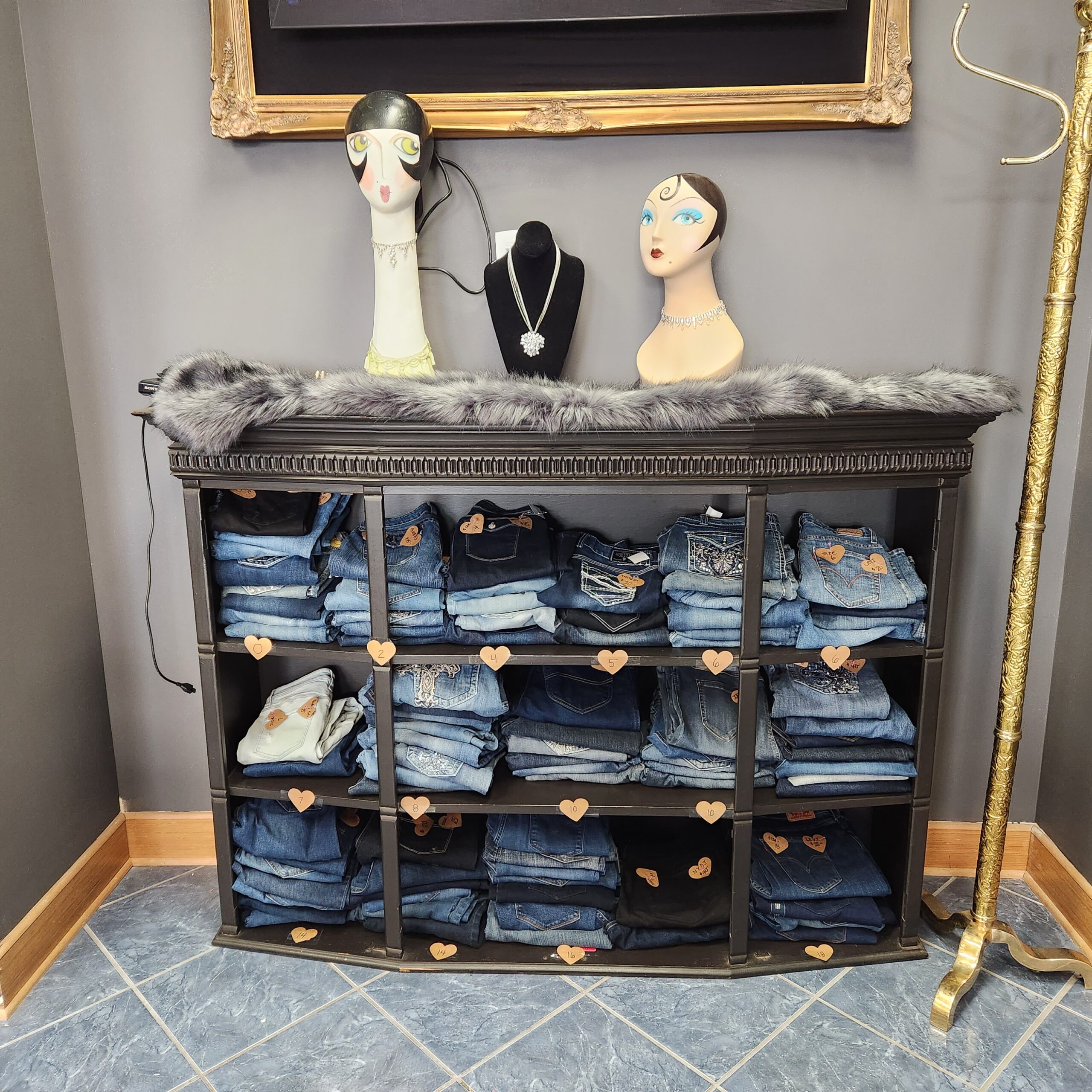 A display shelf holds neatly folded denim jeans, organized in various compartments adorned with heart decorations. Atop the shelf, two mannequin busts adorned with jewelry are displayed alongside a black bust with a necklace. The backdrop is a framed picture.