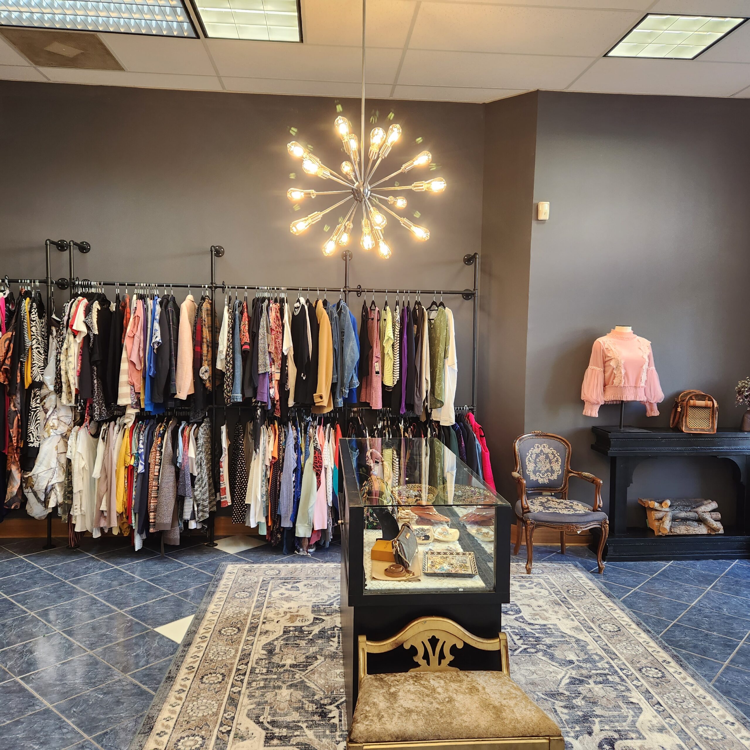 A boutique with a modern chandelier, clothing racks filled with various garments, a glass display table with accessories, an upholstered chair, a black mantel with decor items, and a mannequin wearing a pink blouse. The flooring is blue tiles with a rug.