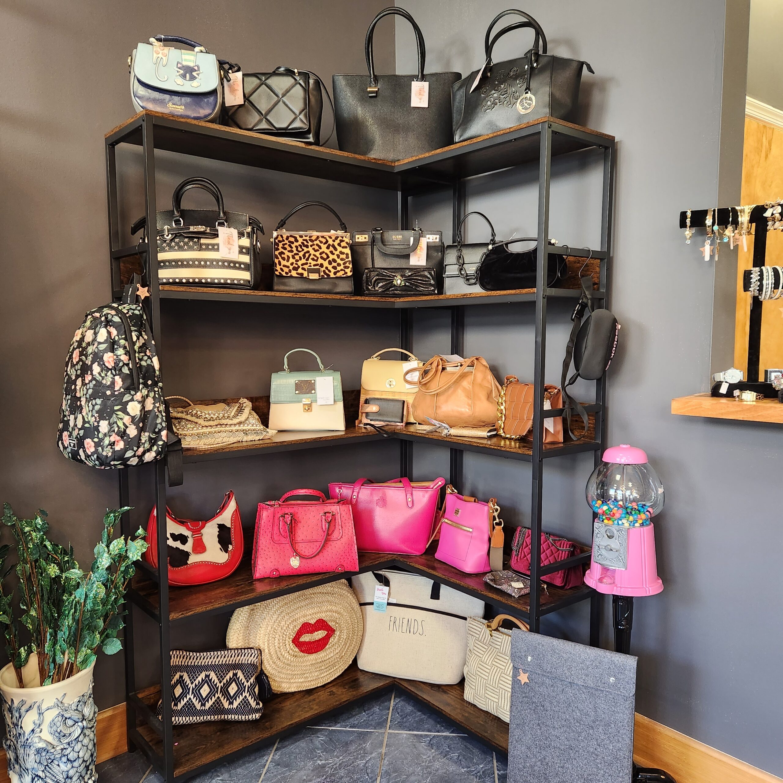 A corner display shelf in a store showcases various handbags in different sizes, colors, and styles, including leopard print and bright pink. At the bottom, there are woven bags. To the right, a gumball machine is filled with colorful gumballs.