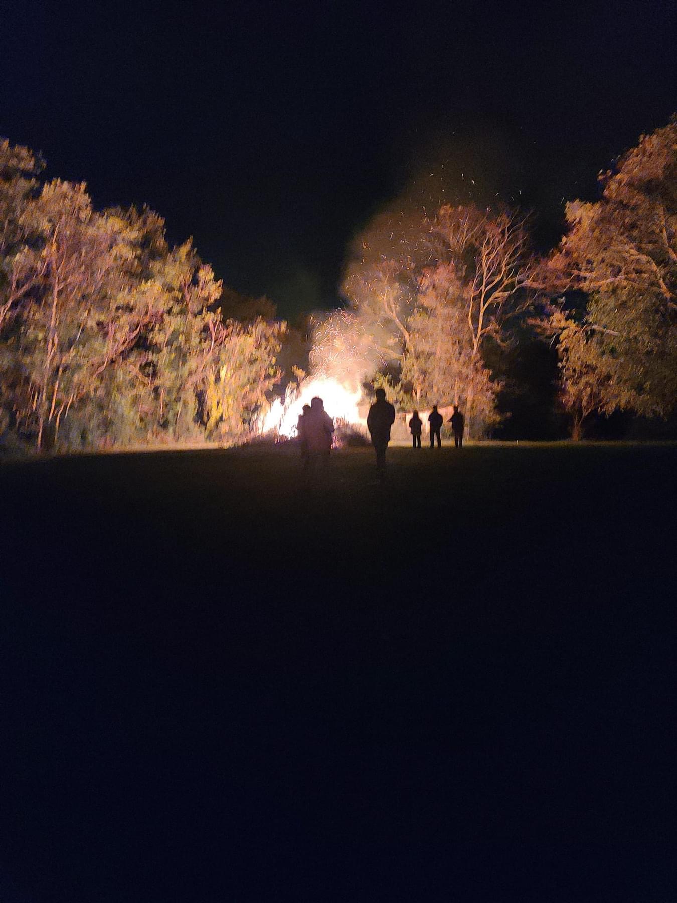 A group of people is seen in silhouette at night, walking towards a large bonfire in the background surrounded by trees. The glowing flames illuminate the dark sky and cast a warm light on the nearby foliage. The scene suggests a gathering or outdoor event.