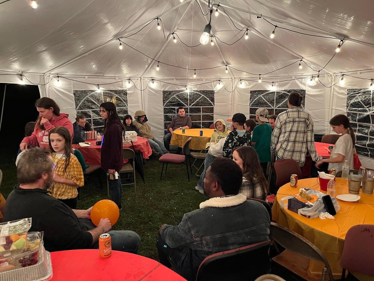 A group of people sit and stand under a large, well-lit, white tent. They are gathered around several round tables covered with orange and yellow tablecloths. Some people are eating or drinking, while others are talking. String lights hang from the tent's ceiling.