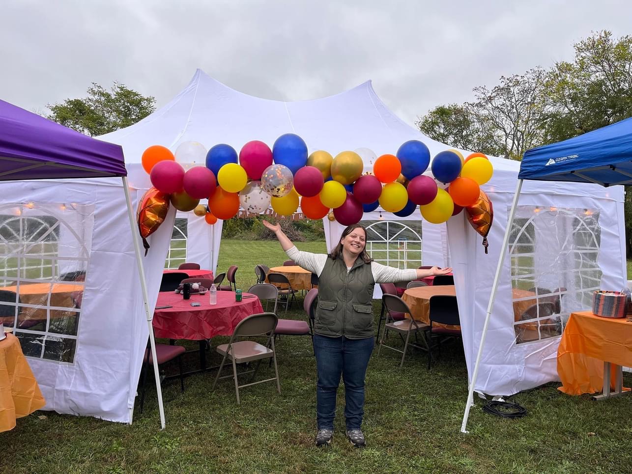 A person stands smiling with arms outstretched in front of a white canopy tent decorated with colorful balloons. Inside the tent, tables have red and orange tablecloths with chairs arranged around them. It's an outdoor setting with a cloudy sky and green grass.