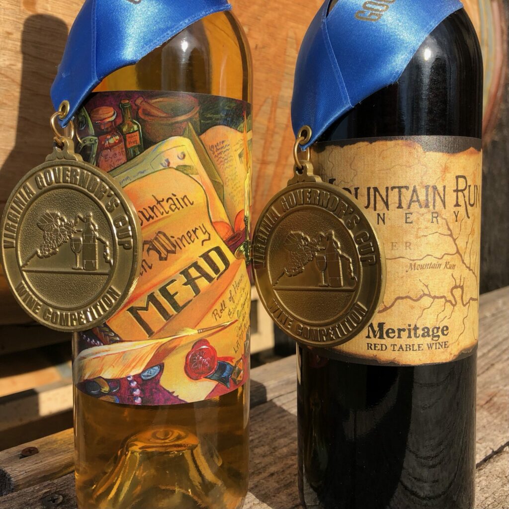 Two wine bottles with blue ribbons and gold medals displayed. The bottle on the left has a colorful label that reads 'Mead,' while the bottle on the right has a rustic label that reads 'Meritage Red Table Wine.' Both medals, from prestigious breweries and wineries, read 'Virginia Governor's Cup Wine Competition.