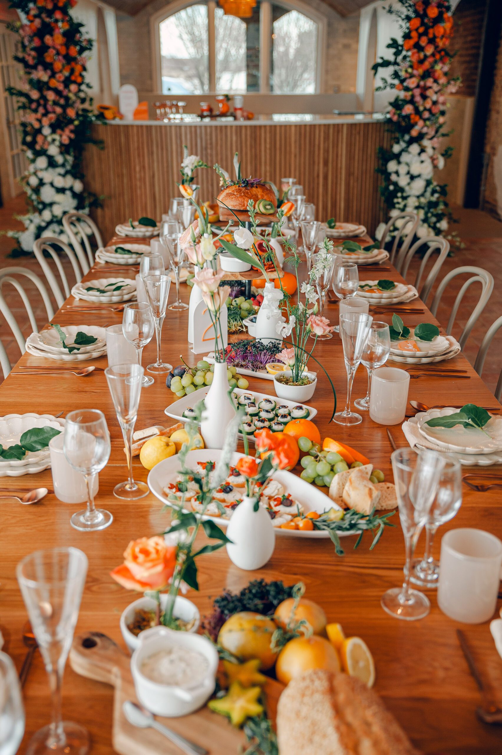 A long wooden dining table is elegantly set with plates, cutlery, and glasses. It features floral centerpieces, greenery, and a variety of food items including bread, fruits, and desserts. The backdrop includes large floral arrangements and a wooden partition.