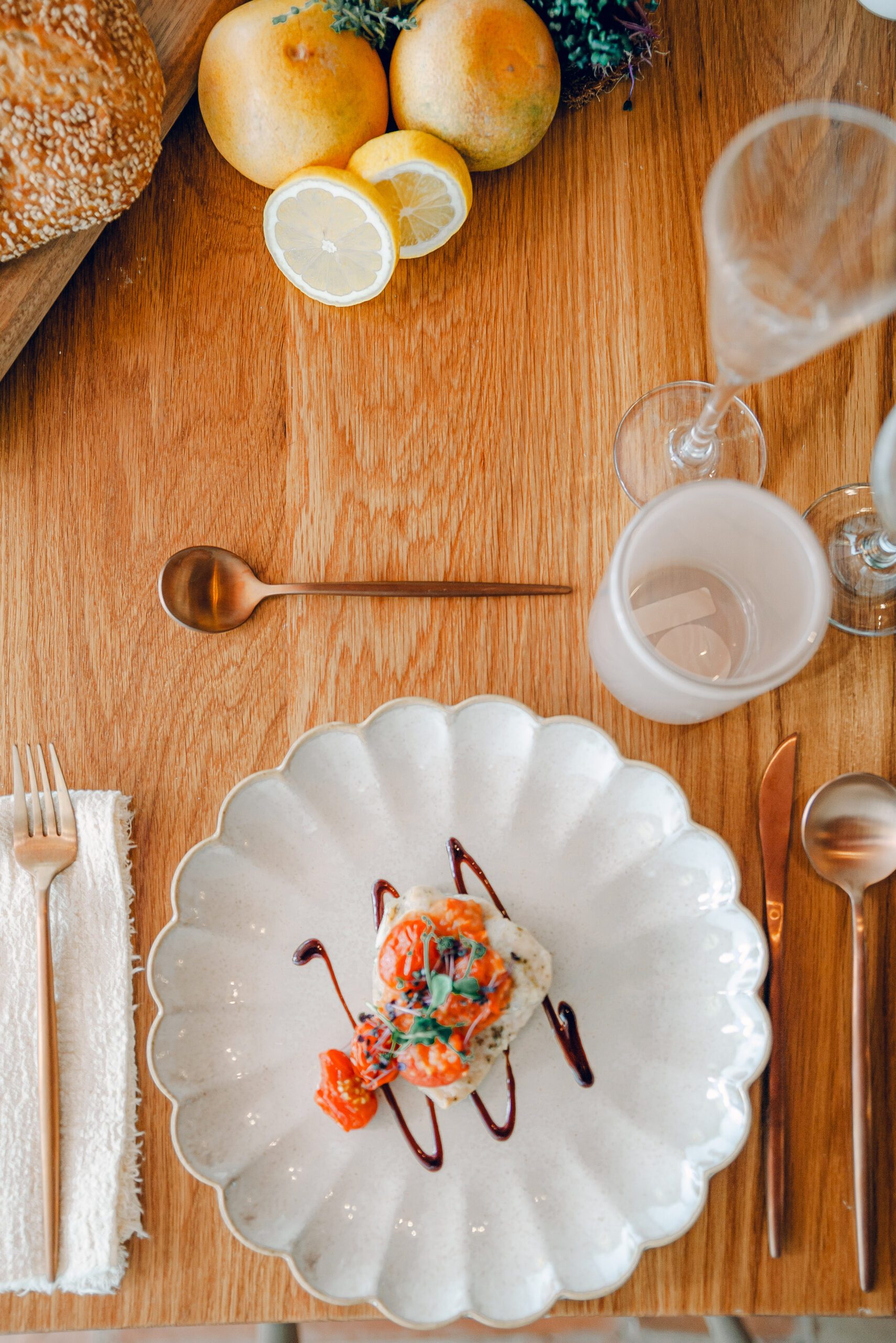 A beautifully plated dish on a white scalloped plate, featuring a gourmet appetizer garnished with herbs and sauce. The wooden table setting includes gold utensils, a white napkin, a glass of water, a champagne flute, and lemons alongside bread in the background.