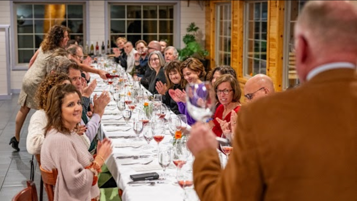 A large group of people sitting around a long, elegant dinner table decorated with flowers and wine glasses. The person at the head of the table holds up a wine glass for a toast, and guests are clapping and smiling, creating a cheerful atmosphere.