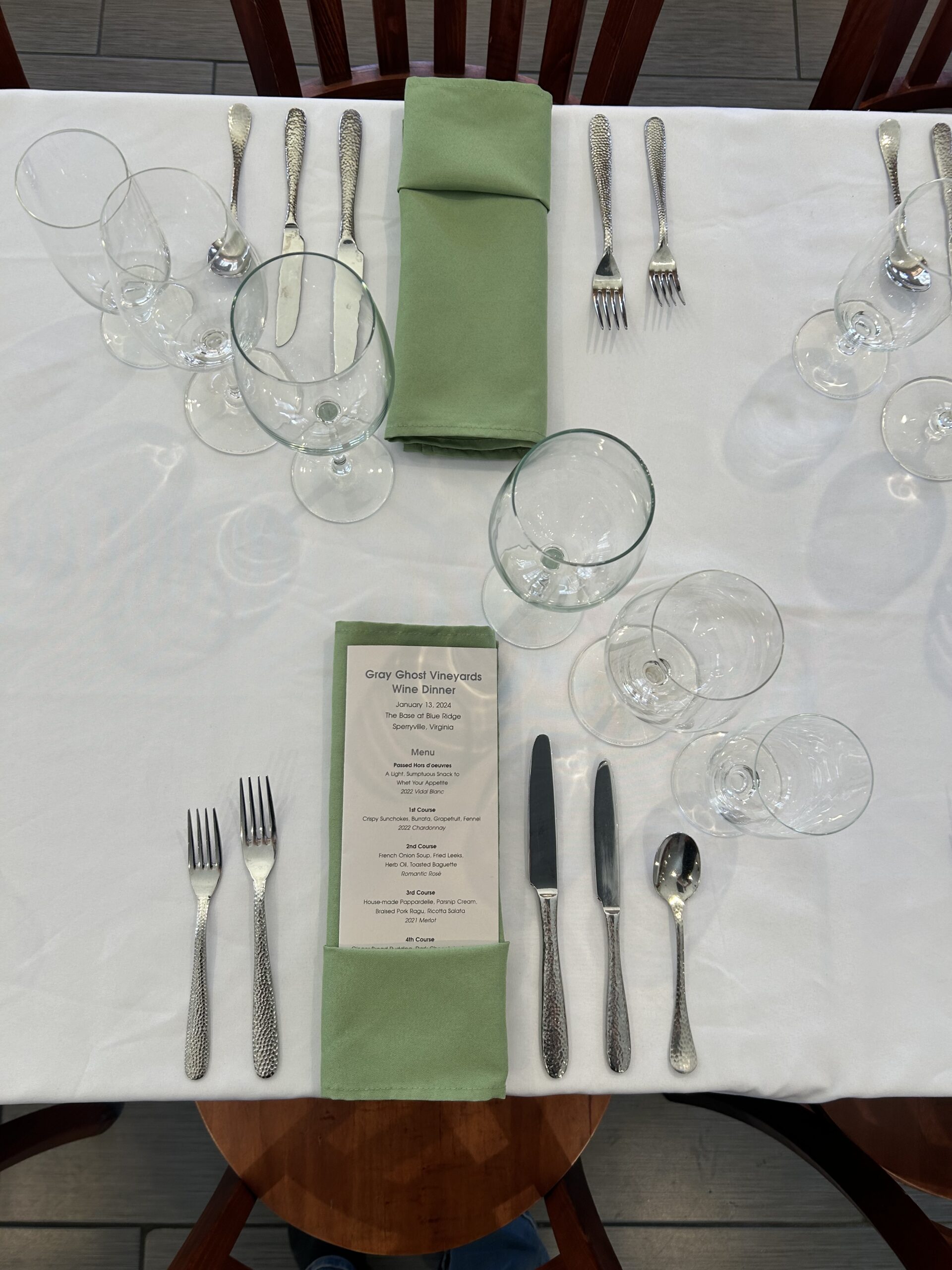 A table set for a formal meal with two place settings. Each setting includes silverware, a green folded napkin, and multiple glasses. A menu titled 