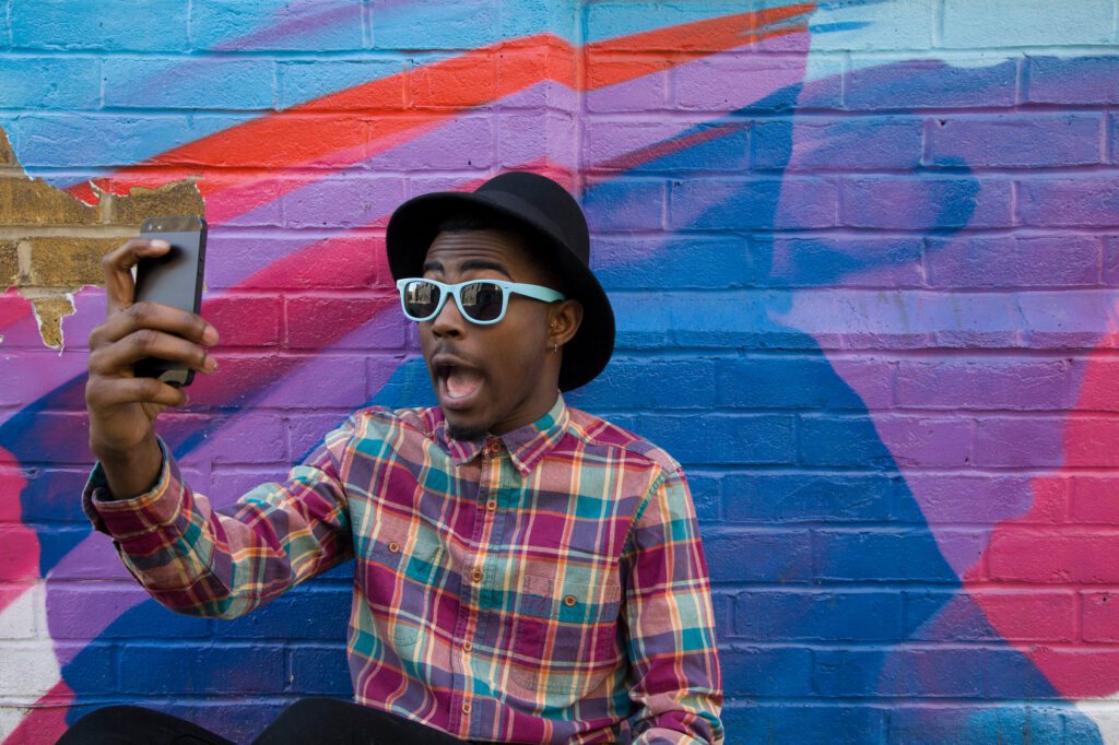 A person wearing a black hat, white sunglasses, and a plaid shirt takes a selfie against a colorful mural. The individual has an open-mouthed, surprised expression as the wall features abstract shapes in blue, red, and purple hues.