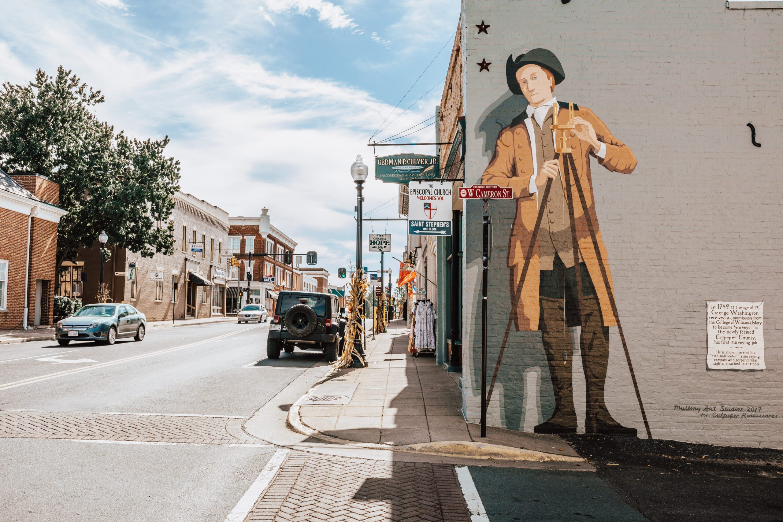 Street view of a small town on a sunny day featuring a large mural of a historical figure on the side of a building. The street has signs for East King Street, colorful banners, lamp posts, and a parked car. Other buildings and trees line the background, adding charm to this picturesque scene.