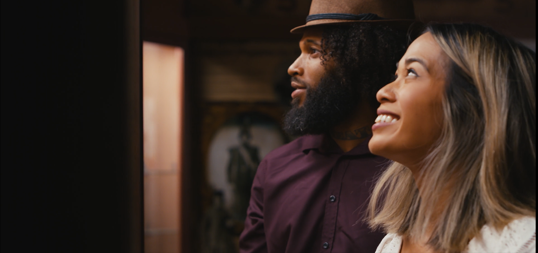 A man with curly hair and a beard wearing a brown hat and a woman with shoulder-length hair smiling look towards the left. Both appear to be inside a warmly lit room. The background is slightly blurred with a glimpse of a framed picture.