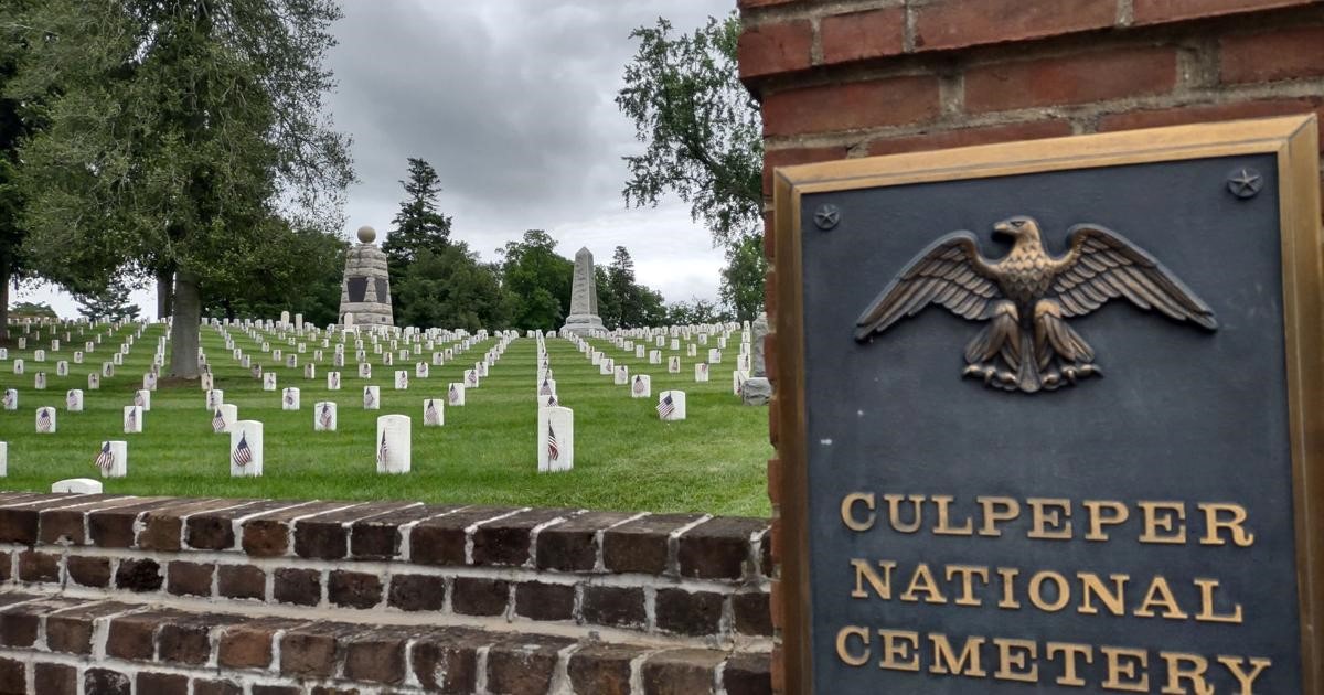 Culpeper National Cemetery, with a close-up of a plaque featuring an eagle and the cemetery name on a brick wall in the foreground. Rows of white headstones and various memorials are visible under a cloudy sky. Trees dot the well-maintained grounds.