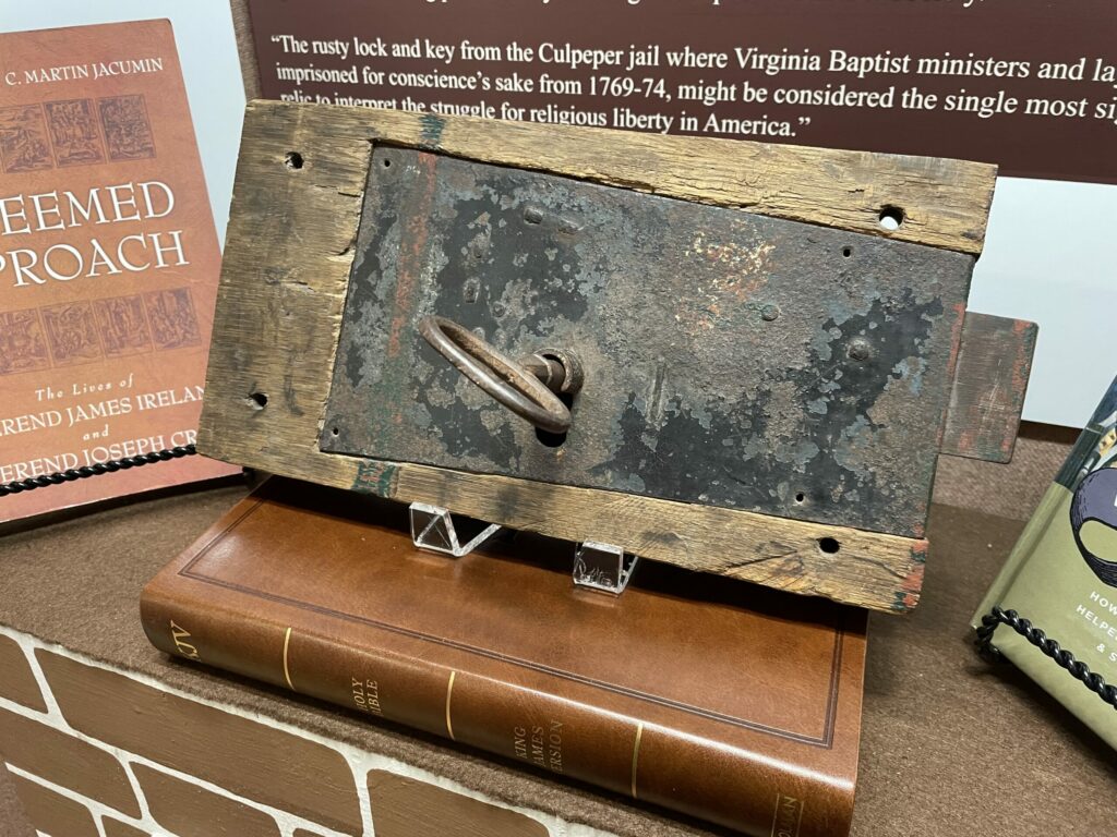 A close-up of an old, rusted lock and key mounted on a wooden board displayed in the museum exhibit. The lock is weathered, indicating age. The display is set against a backdrop with historical text and books related to Baptist ministers and religious liberty in America.