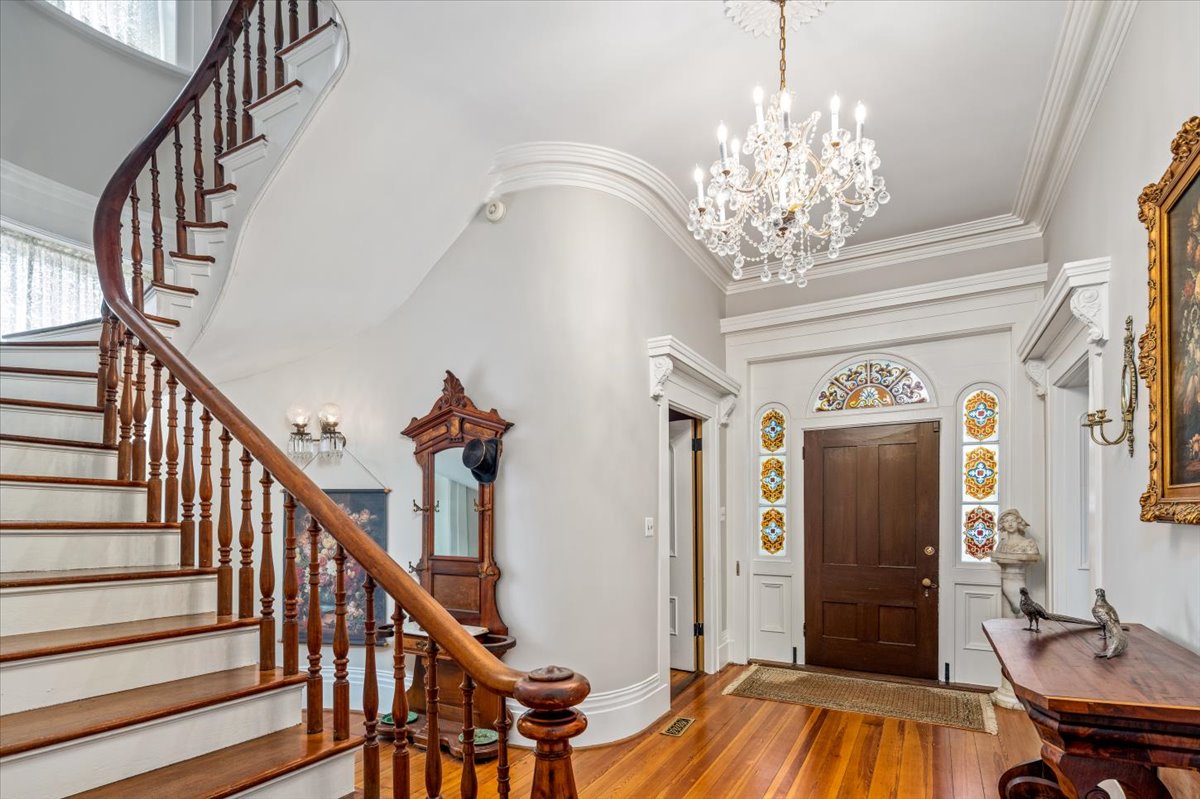 A luxurious entryway at Hill Mansion features a grand wooden staircase with ornate railings. The front door is adorned with colorful stained glass panels. A chandelier hangs from a high ceiling, illuminating polished wood floors and antique furniture, including a mirror and side table.