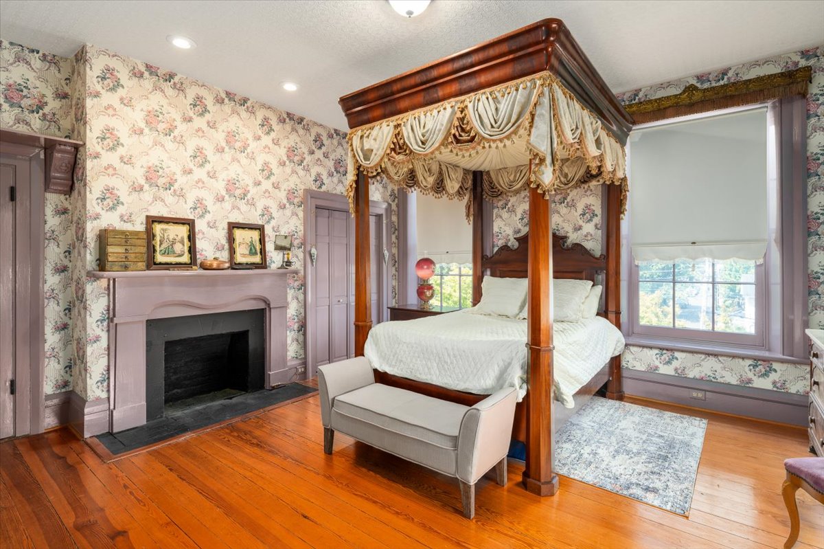 A cozy, vintage-style bedroom at Hill Mansion features a four-poster bed with draped canopies, floral wallpaper, hardwood floors, and a decorative fireplace. Two framed pictures adorn the wall above the fireplace. A small, cushioned bench sits at the foot of the bed.