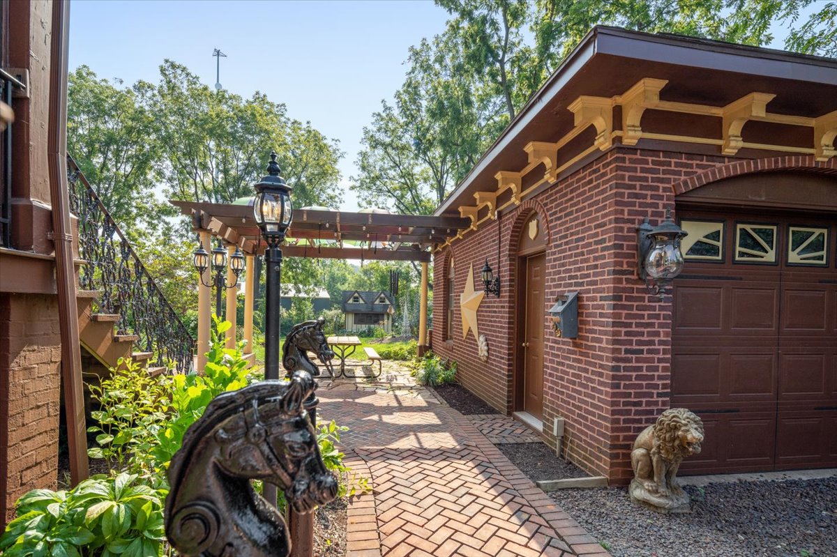 A brick pathway lined with plants and horsehead posts leads to Hill Mansion's brick garage with a brown door and star decoration. A pergola with lanterns stands beside the garage, and a lion statue is near the pathway. Tall trees and a decked seating area with chairs are in the background.