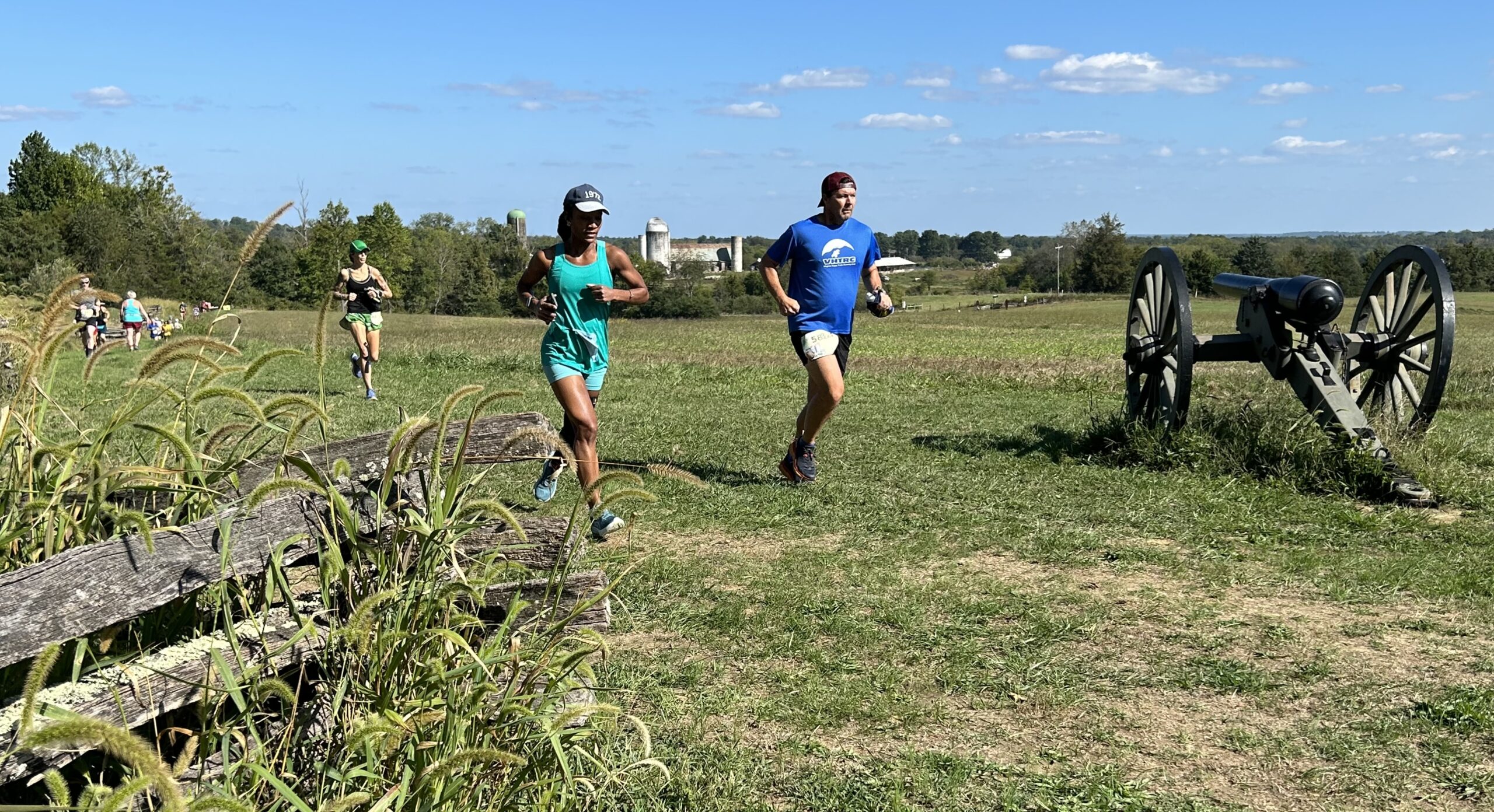 Runners in athletic gear participate in a race on a grassy field with clear blue skies. The scene includes an old cannon on the right and more racers in the background. Wooden fencing and tall grasses are in the foreground.