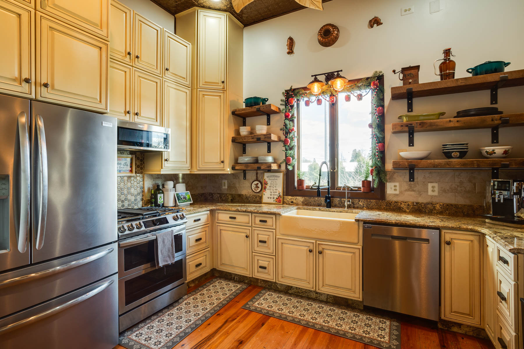 A cozy kitchen at Hill Mansion features cream cabinets, granite countertops, a farmhouse sink below a window decorated with colorful ornaments, stainless steel appliances, open shelving with neatly arranged dishes, and warm wooden floors. A patterned rug runs across the floor.