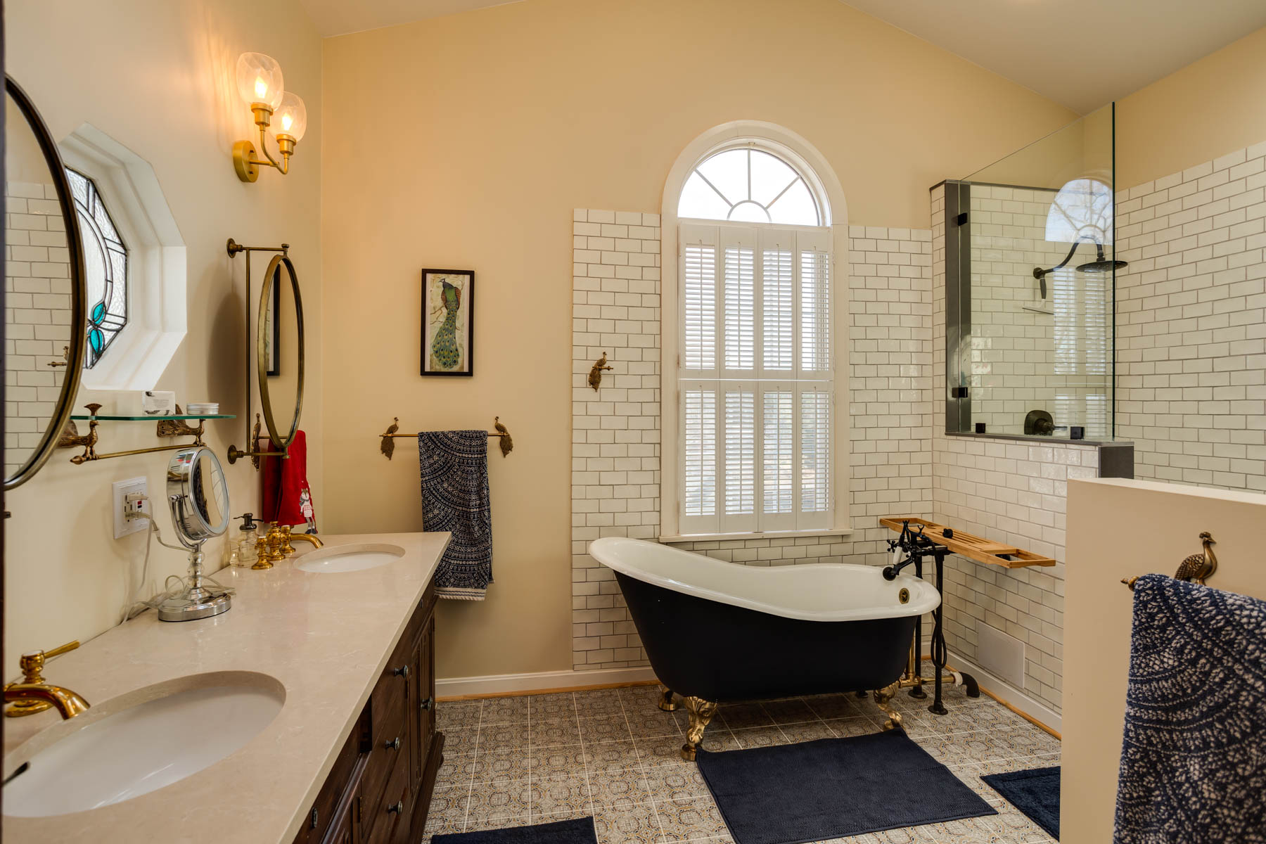 The Hill Mansion bathroom features a black clawfoot bathtub with a wooden tray, double sinks on a wooden cabinet, round mirrors, white tiled walls, and a large arched window with shutters. Decorative wall lights and blue towels add to the cozy ambiance.