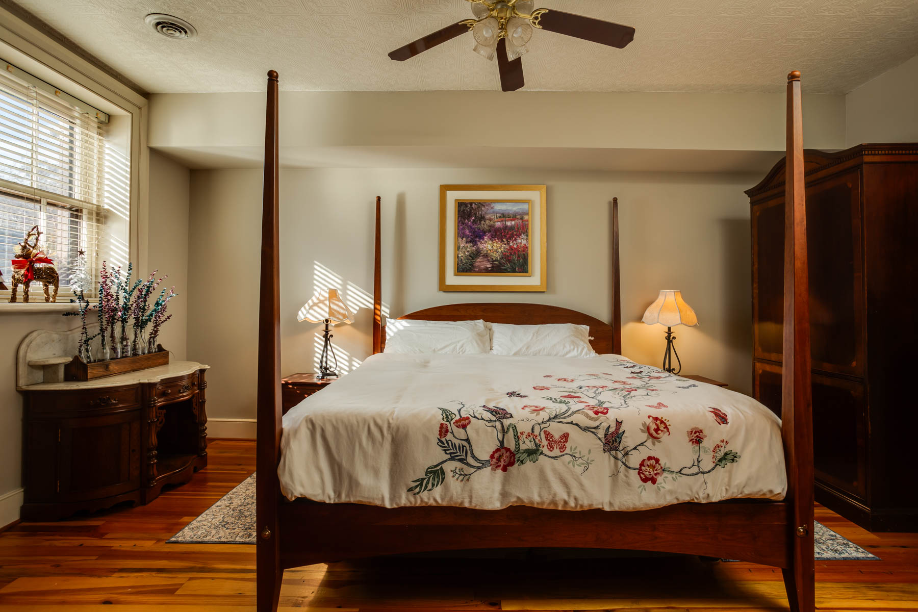 A cozy bedroom in Hill Mansion features a wooden four-poster bed with a floral embroidered white quilt. Two bedside tables hold lamps with white shades. An armoire stands to the right, and a colorful painting hangs above the bed. Blinds cover a window on the left.