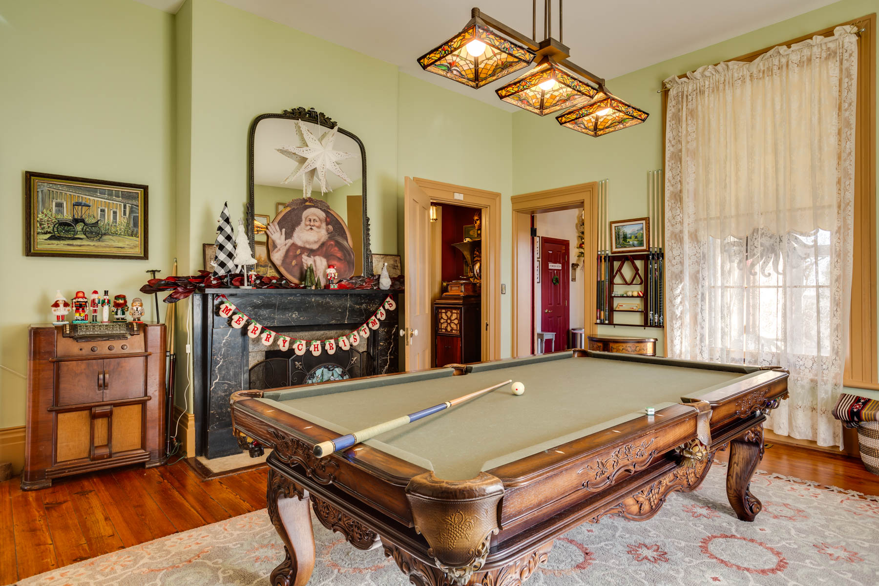 The Hill Mansion features a room with a pool table at the center, decorated with Christmas ornaments. The walls are light green, and a large mirror above the fireplace reflects a Santa Claus picture. There is a lace curtain on the window, wood flooring, and vintage-style lighting above the table.