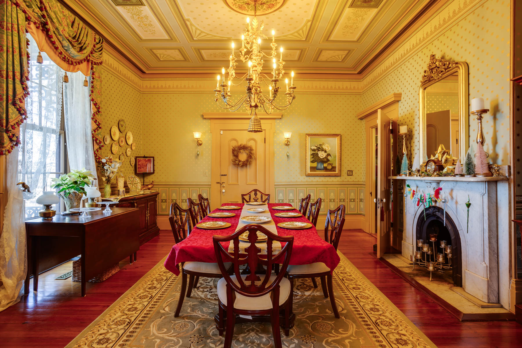 A grand dining room in Hill Mansion boasts an ornate chandelier hanging over a long table set for dinner. The table is adorned with a red tablecloth and gold-rimmed plates. The room features elegant details, including a decorative fireplace, wall patterns, and large windows with luxurious drapes.