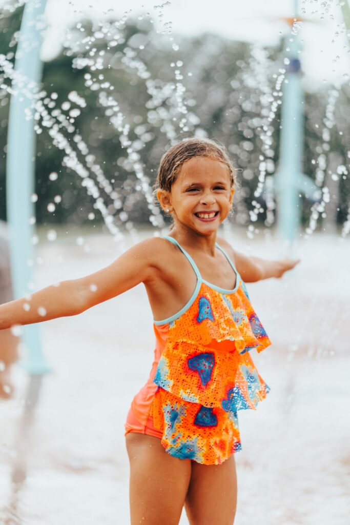 A young girl with short hair is wearing an orange swimsuit with blue and pink patterns. She is smiling broadly with her arms outstretched, standing under a cascading water fountain, enjoying the free fun in Culpeper on a sunny day. Trees are blurred in the background.