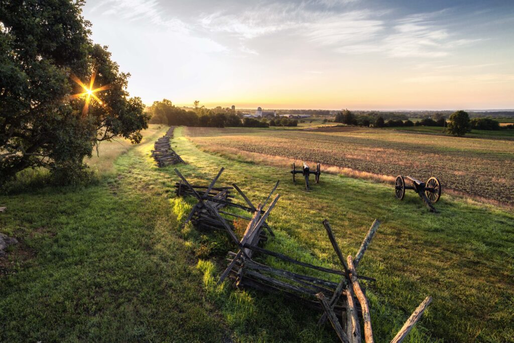 A scenic countryside landscape at sunrise, featuring a split-rail fence leading into the distance across grassy fields. Historical cannons are positioned on either side of the fence line, with a clear sky and rising sun casting a warm glow over the scene.