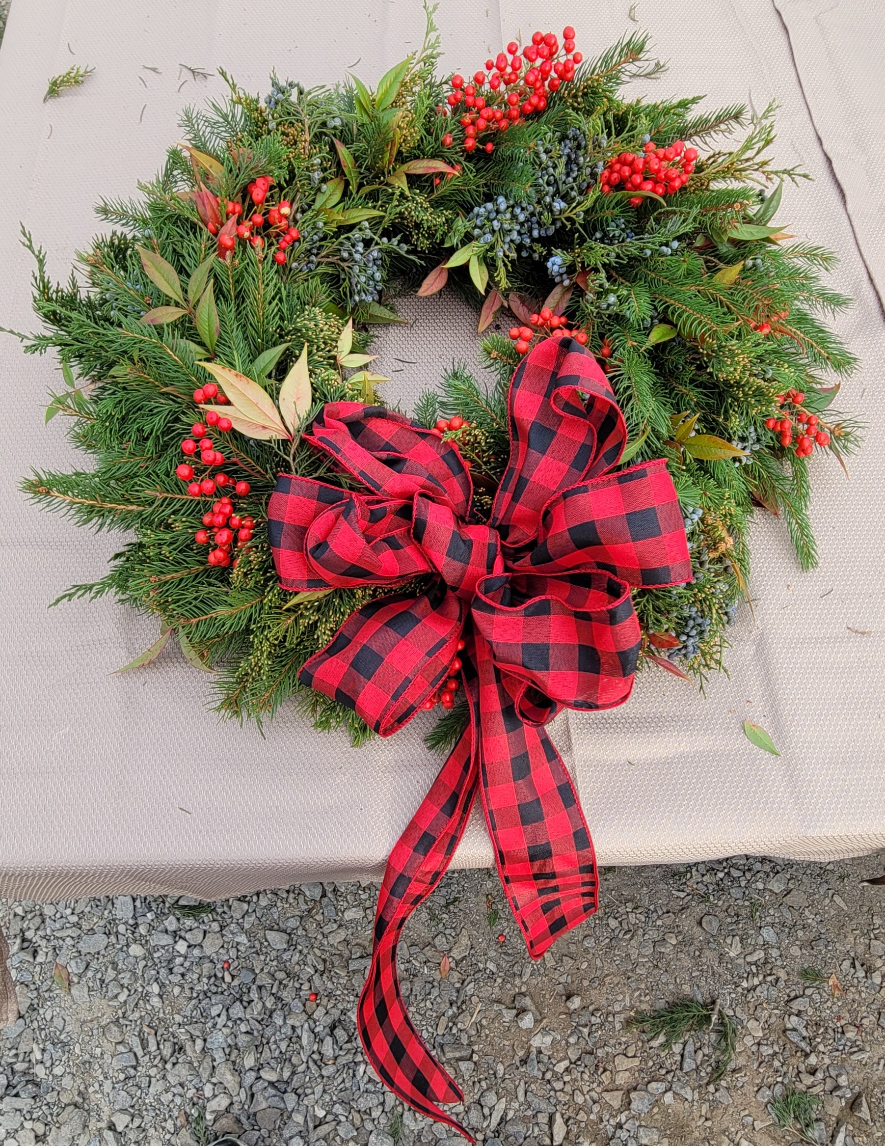 A festive holiday wreath made of evergreen branches, clustered with berries, and adorned with a large red and black plaid bow, is displayed on a light-colored cloth surface with gravel seen in the background.