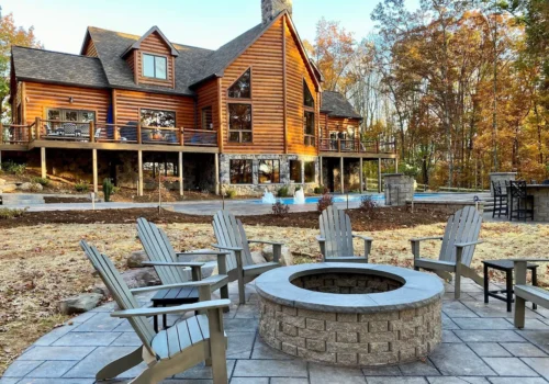 A large log cabin on Lake Culpeper with multiple windows, balconies, and a stone chimney stands amidst autumn trees. In the foreground, a circular fire pit area is surrounded by six wooden chairs on a paved patio. A swimming pool and outdoor seating area are visible near the house. Image