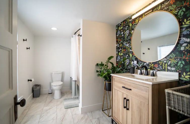 A modern bathroom features a round mirror above a wooden vanity with a sink, faucet, and potted plant. The wall behind the vanity has a colorful, botanical wallpaper. To the left, there's a toilet and a shower with a white curtain. The floor is light tiled.