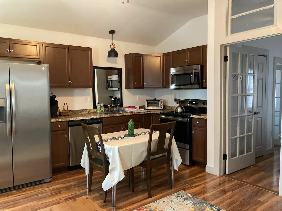 A modern kitchen with wooden cabinets, stainless steel appliances, and granite countertops. A small dining table with a white tablecloth and two chairs is placed in the center. A French door leads to another room. The floor is wooden and there is a rug near the table.