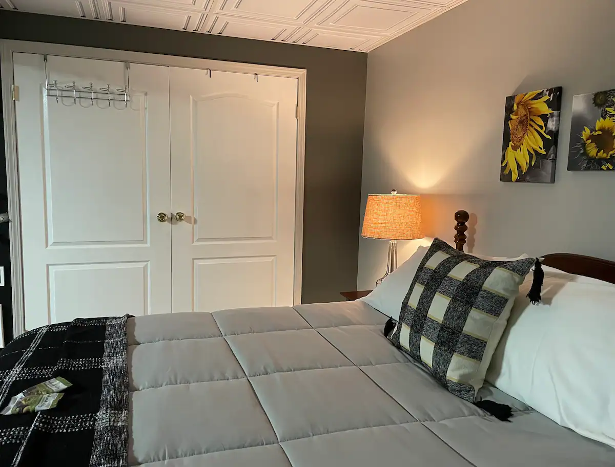 A cozy bedroom with a neatly made bed featuring a gray comforter and black-and-white checkered pillows. A bedside lamp casts a warm light beside sunflower-themed wall art. Double white doors with clothes hooks are visible in the background.