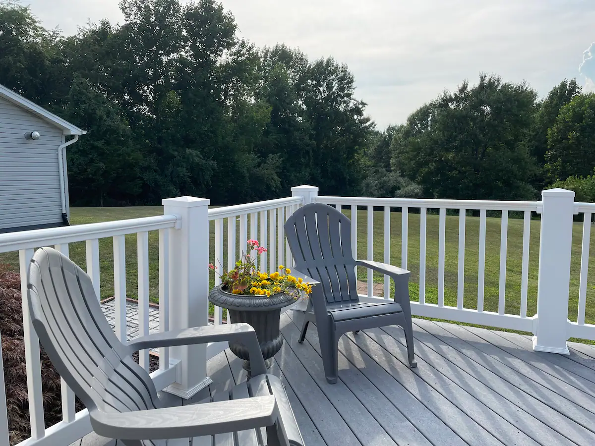 A spacious deck with two gray Adirondack chairs and a planter with colorful flowers. The deck has a white railing and overlooks a grassy yard with dense trees in the background under a partially cloudy sky.