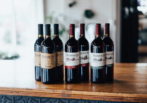 Five bottles of wine with various labels are displayed on a wooden table indoors. Image