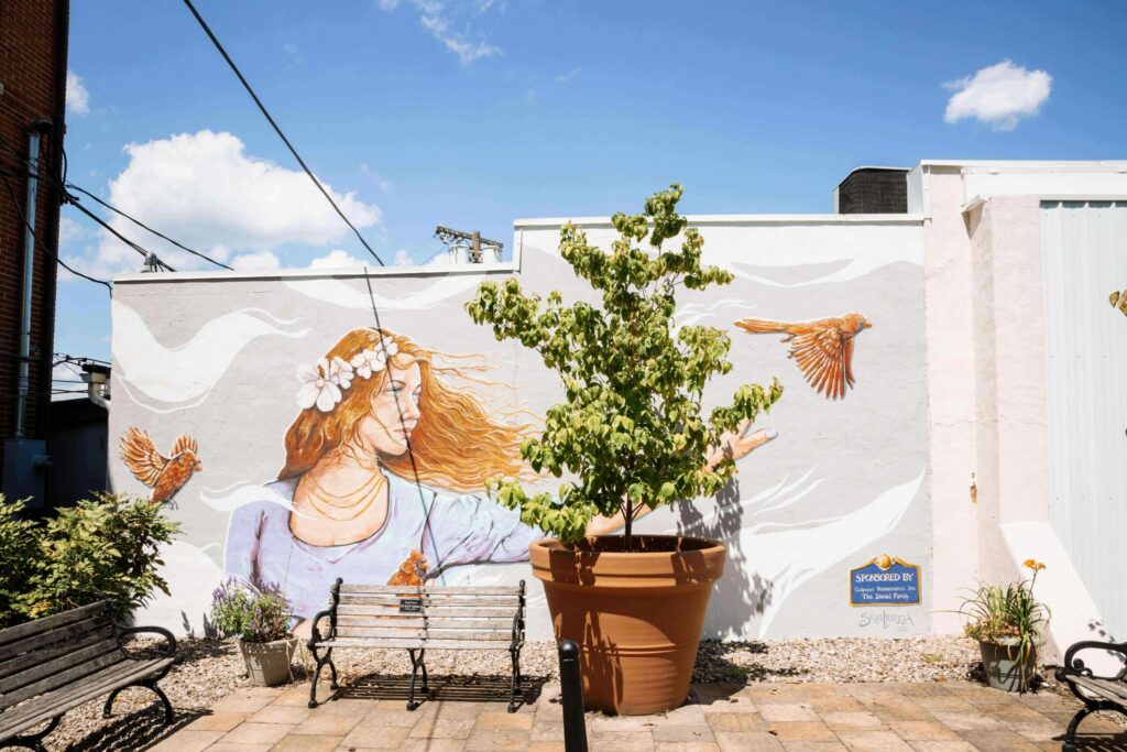 goddess mural painted on a wall in a park with hair blowing in the wind and a tree and bench in front