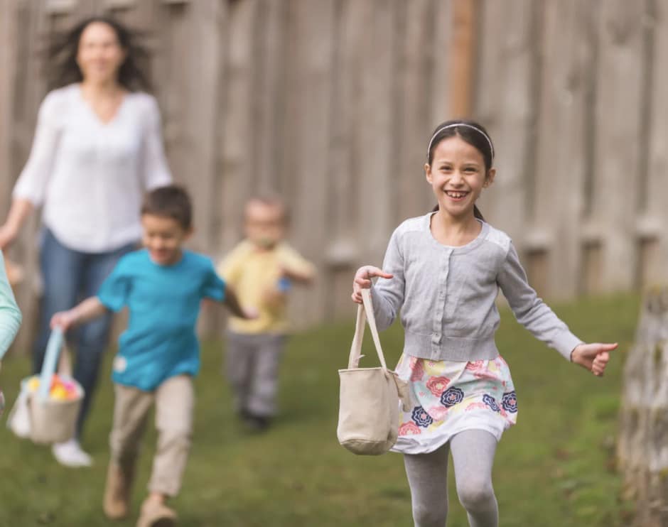 three children and an adult running through the grass with Easter baskets in their hands, looking for Easter eggs.