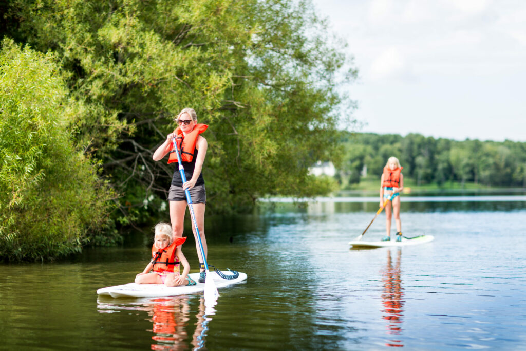 women standing on a paddle board in a lake with a child sitting and another child standing on a paddleboard behind her