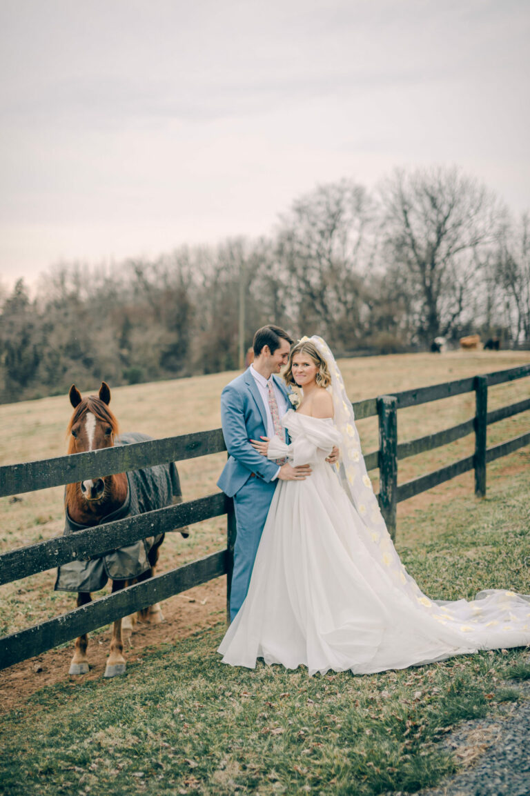 couple in wedding attire standing next to a fence with a horse behind them