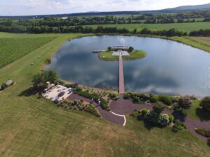 An aerial view of a serene pond with a wooden walkway leading to a small island gazebo. Surrounding the pond are lush green fields and grass. Near the pond's edge is a paved walkway and landscaped garden area with a seating space.