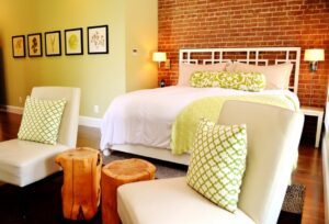 A modern bedroom with a white bed frame, green and white bedding, and matching accent pillows. The room has a brick accent wall, five nature-themed framed pictures, two cream-colored chairs with patterned cushions, and two wooden stump side tables.